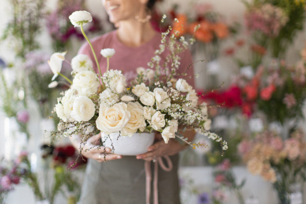 A florist carefully holding an elegant ivory floral centerpiece, with vibrant, colorful blooms artistically blurred in the background, creating a soft, romantic aesthetic.