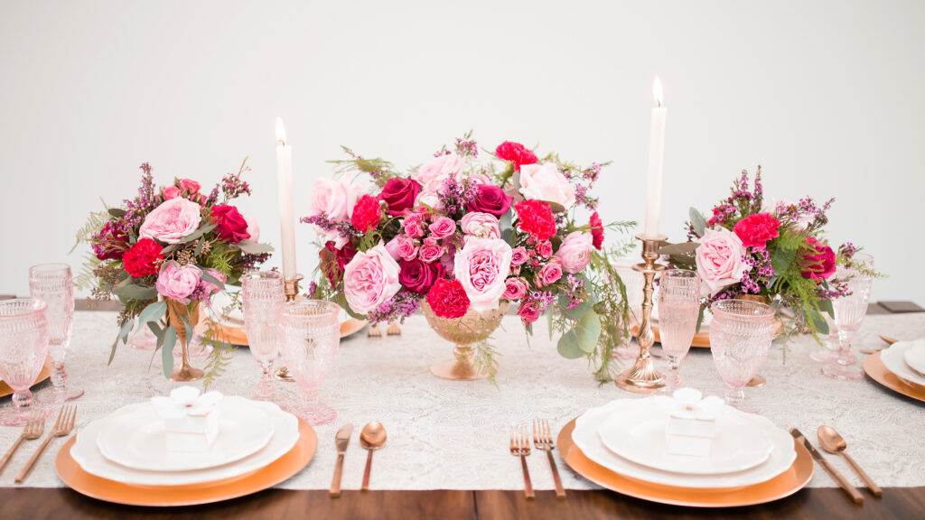 A romantic tablescape featuring three elegant centerpieces with blush and fuchsia blooms, surrounded by pink-hued glassware. The arrangement is set on a soft linen tablecloth, creating a cohesive and luxurious wedding design.