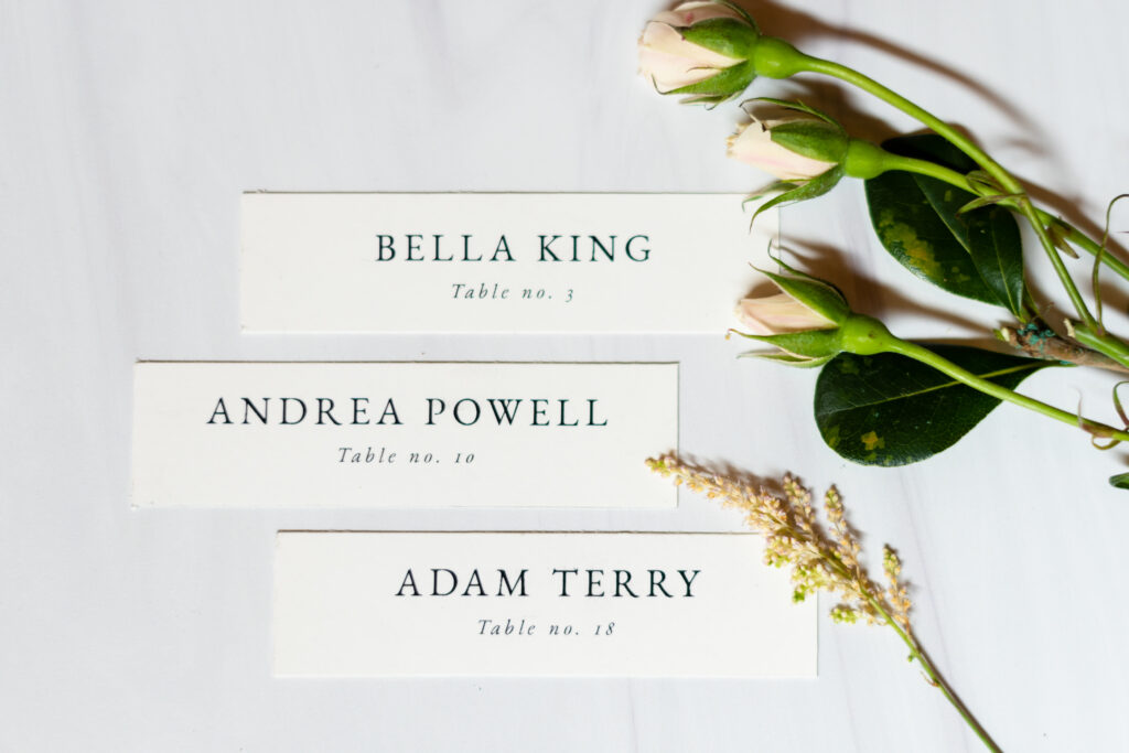 Three elegantly designed name tags with black and white typography, laying flat on a table surrounded by delicate greenery, creating a minimalist and refined aesthetic.