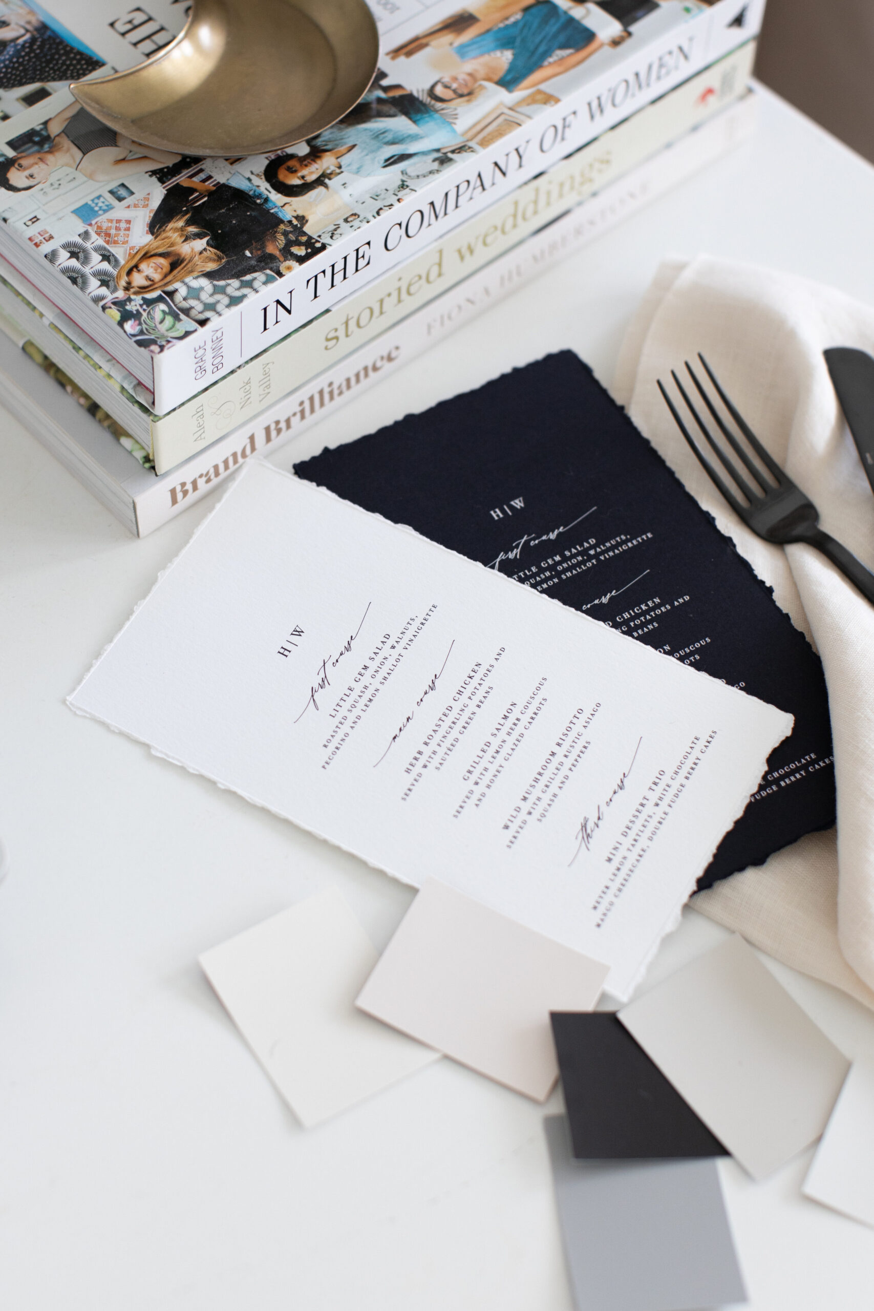 Elegant black-and-white wedding stationery styled alongside fabric swatches, gleaming black cutlery, and a curated stack of books.